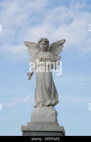 Steinerne Engelsstatue mit Blumen auf dem Friedhof in Taormina, Sizilien, Italien, vor einem blauen und bewölkten Himmel. Stockfoto