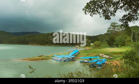 Kleine Boote, die am grünen Ufer des Tuyen-Lam-Sees vor Anker liegen, umgeben von Kiefernwaldhügeln unter einem dramatischen stürmischen Himmel in Da Lat, Vietnam. Stockfoto