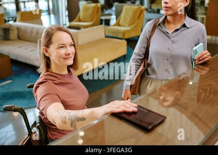 Frau mit Behinderung checkt an der Rezeption eines modernen Hotels ein Stockfoto