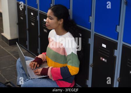 Mädchen in gestreiftem Pullover und Jeans sitzt im Schulflur und verwendet einen Laptop neben blauen Spinden Stockfoto