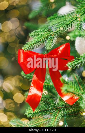 Hellrote Schleifenbänder schmücken einen grünen Weihnachtsbaum mit weißen Ornamenten und warmen goldenen Bokeh-Lichtern im Hintergrund Stockfoto