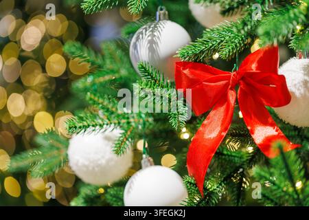 Hellrote Schleifenbänder schmücken einen grünen Weihnachtsbaum mit weißen Ornamenten und warmen goldenen Bokeh-Lichtern im Hintergrund Stockfoto