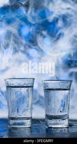 Zwei gekühlte Wodka-Shots in nassen Gläsern mit Kondenswasser, umgeben von wirbelndem weißem Rauch und atmosphärischem blauem Licht. Dramatische vertikale Komposition Stockfoto