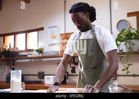 Afrikanischer Mann rollt Teig in der Küche, trägt eine grüne Schürze und ein weißes T-Shirt, verwendet einen Nudelholz Stockfoto