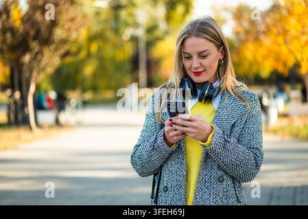 Schöne und elegante Geschäftsfrau trägt kabellose Kopfhörer und tippt auf ihrem Smartphone, während sie an einem Herbsttag im Stadtpark steht. Stockfoto