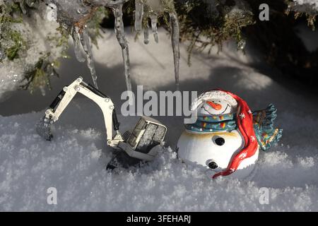 Spielzeuggraue Mini-Bagger und ein Souvenir-Schneemann im Schnee inmitten gefrorener Natur an einem sonnigen Wintertag. Stockfoto