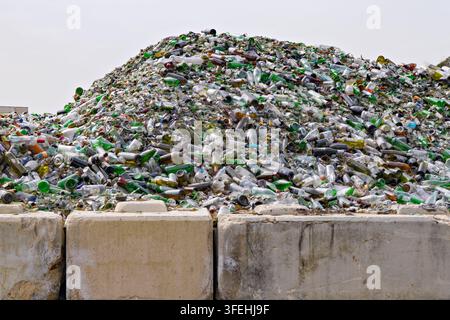 Riesiger Haufen weggeworfener Glasflaschen und zerbrochener Behälter hinter Betonbarrieren in der Recyclinganlage, der die Abfallansammlung und Materialrecycling zeigt Stockfoto