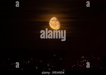 Der leuchtende Mond im abnehmenden Vollmond steht hell durch verschwommene Wolken über dem fernen Stadtlicht in der Nacht Stockfoto