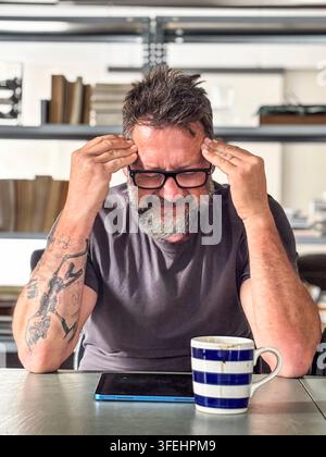 Bartmann mit Brille reibt sich frustriert die Schläfen, während er an einem Heimarbeitsplatz mit einem Tablet und einer gestreiften Kaffeetasse sitzt, und vermittelt Stress, Stockfoto