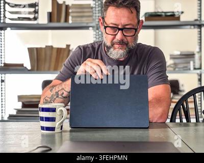 Bartträger mit Brille arbeitet an einem Laptop an einem Tisch in einem lässigen Heimbüro, Kaffeetasse in der Nähe und Regale mit Büchern und Akten im Hintergrund c Stockfoto