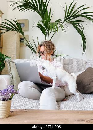 Frau mit lockigem Haar und Brille, die remote auf einem Laptop arbeitet, während ihr weißer Hund neben ihr auf der Couch steht und eine gemütliche und komfortable Atmosphäre schafft Stockfoto