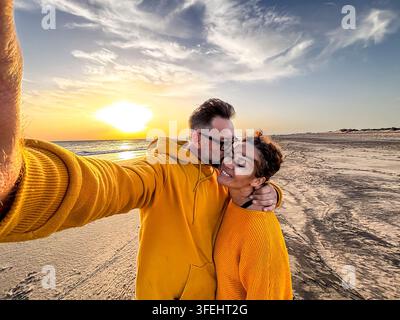 Paar in passenden gelben Pullovern teilt einen liebevollen Selfie-Kuss an einem Sonnenuntergangsstrand, umarmt Freude und Wärme, während die Wellen funkeln, und fängt romantische Verbindung ein Stockfoto