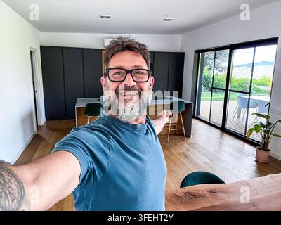 Glücklicher Mann, der ein Selfie in seinem neuen modernen Haus macht, präsentiert die geräumige, offene Küche mit einer großen Schiebetür, die sich zum Außenbereich öffnet Stockfoto