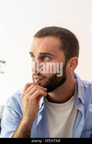 Nachdenklicher bärtiger Mann in blauem Hemd, der in einem lässigen Porträt weg schaut, nachdenklicher Ausdruck und entspannte Pose Stockfoto