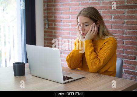 Junge Frau in einem gelben Pullover sitzt an einem Holztisch und verwendet einen Laptop in einem hellen Heimbüro mit einer Ziegelwand, wirkt nachdenklich und leicht bor Stockfoto
