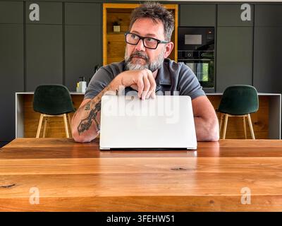 Mittelalterer bärtiger Mann arbeitet an einem Laptop an einem Holztisch in der Küche und blickt nachdenklich weg. Lässige Home-Office-Stimmung mit Tattoos, Brille, modernem i Stockfoto