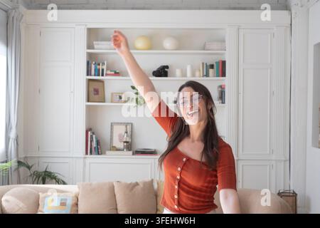 Frau mit Brille lächelt und lacht, während sie fröhlich tanzt, einen Arm in einem häuslichen Innenraum hebt und Freude, Freiheit, Energie und Feierlichkeit ausdrückt Stockfoto