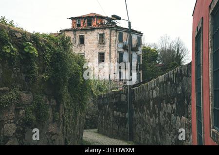 schaut eine kurvenreiche Kopfsteinpflastergasse hinunter, die von hohen, moosbewachsenen Steinmauern eingeschlossen ist. Im Hintergrund steht ein großes, stark verfallenes historisches Gebäude Stockfoto