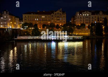 Nachtansicht über die Moldau mit einem beleuchteten Ausflugsschiff und historischen Gebäuden entlang der Uferpromenade. Stockfoto