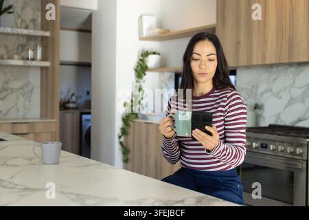 Frau sitzt an Kücheninsel, hält grüne Tasse und überprüft Smartphone, gestreiftes Hemd Stockfoto
