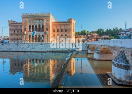 Stadt hall von Sarajevo und Seher-?ehaja-Brücke, die sich im Fluss Miljacka spiegelt, mit traditionellen Hügelhäusern im Morgenlicht, Sarajevo, Sarajevo Kanton Stockfoto