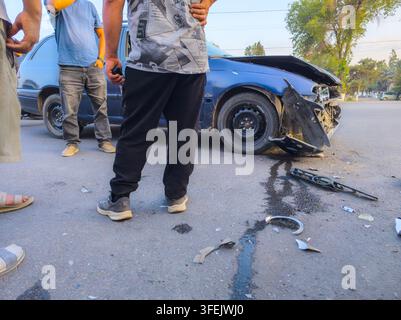 Eine Gruppe von Männern versammelt sich um ein schwer beschädigtes blaues Auto auf einer asphaltierten Straße. Die Front ist eingedrückt, mit verstreuten Trümmern und einer dunklen Flüssigkeit, die auf den Asphalt tropft. Stockfoto