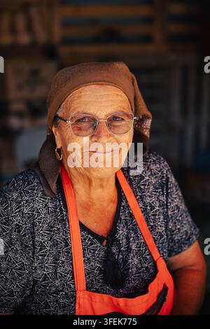 Senior-Frau, Verkäuferin, die eine Brille und ein Kopftuch trägt, lächelt als örtliche Markthändlerin hinter ihrem Gemüsestand auf einem Bauernmarkt. Stockfoto