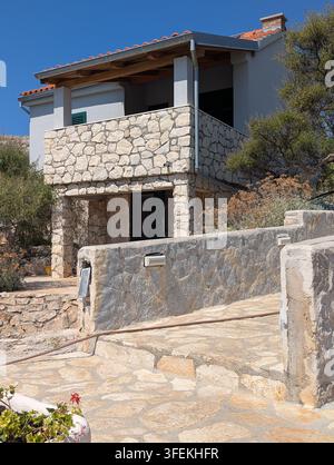 Pirovac, Kroatien, 16. August 2026: Sonnenbeschienene Steinvilla mit Balkon, Terrasse, Fensterläden, Veranda und gestuftem Gehweg neben trockenem Landschaftsgarten und klarem Blau Stockfoto