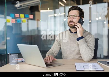 Professioneller Mann führt ein Geschäftsgespräch am Mobiltelefon, nutzt einen Laptop an seinem Schreibtisch in einem modernen Büro, symbolisiert Fernarbeit, Kommunikation und Konnektivität Stockfoto