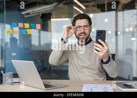 Junger Geschäftsmann mit Brille lächelt und hält ein Smartphone, aktiv an einer virtuellen Besprechung oder Videokonferenz von seinem modernen Büroarbeitsplatz aus teilnehmend Stockfoto