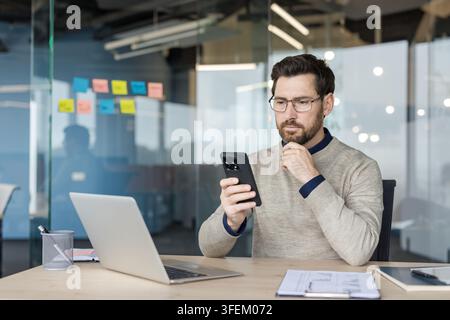 Professioneller Mann mit Brille sitzt an einem Schreibtisch, überprüft Inhalte auf einem Smartphone, denkt über Geschäftsideen in einer modernen Glasbüro-Umgebung nach Stockfoto
