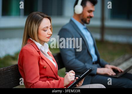 Geschäftsfrau schreibt auf einem Tablet, während ein Mann mit Kopfhörern an einem Laptop arbeitet Stockfoto