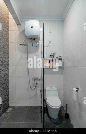 Sauberes modernes Badezimmerinterieur mit einer Glasduschtrennwand, weißer Toilette, wandmontiertem Warmwasserbereiter und hellen Fliesenwänden. Stockfoto