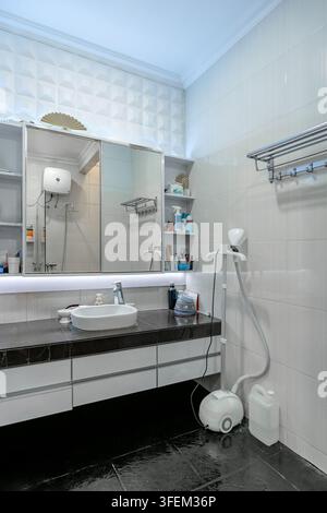 Modernes luxuriöses Badezimmerinterieur mit einem schwarzen Marmorwaschtisch, einem weißen Aufsatzwaschbecken, einem großen beleuchteten Spiegel, strukturierten Akzentfliesen und einem garme Stockfoto