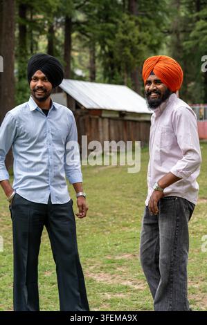 Unbefangener Dreiviertelblick auf zwei junge punjabische Sikh-Freunde, die Turbane tragen, lächeln und wegsehen, während sie im Devidarh-Park stehen. Stockfoto