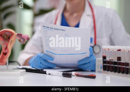 Medizinischer Laborarbeiter, der einen HPV-DNA-Testbericht neben einem Beckenanatomiemodell, blauen Handschuhen, Blutprobenröhrchen und einem klinischen Schreibtisch im hellen Licht überprüft Stockfoto