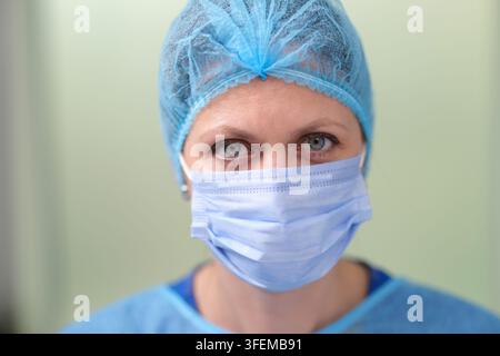 Nahaufnahme-Porträt einer maskierten Gesundheitsarbeiterin mit blauem Chirurgenmütze und Schutzuniform, die direkt in die Kamera blickt mit konzentrierter Ruhe Stockfoto