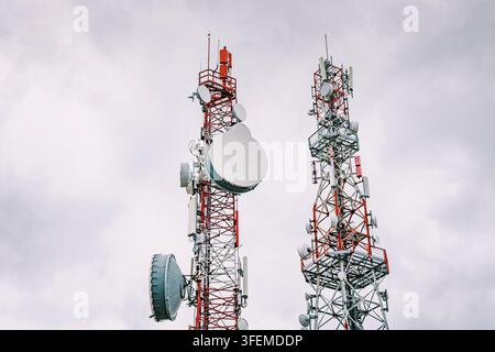 Paar rote und weiße Stahl-Fernkommunikationsmasten, die mit mehreren Satellitenschüsseln und Antennen ausgestattet sind, erheben sich vor einem bewölkten, bedeckten Himmel. Stockfoto