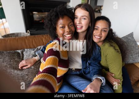 Verschiedene Frauenfreundinnen sitzen auf der Couch, lächeln, machen Selfie mit Smartphone und tragen eine Jeansjacke Stockfoto