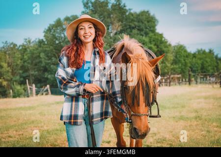 Zufriedene Frau mit Hut, die lächelnd ein Pferd auf einer Wiese auf einer Landranch führt Stockfoto