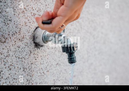 Hand dreht einen Außenhahn aus Metall, der an einer strukturierten Putzwand montiert ist, Wasser fließt frei aus dem Auslauf. Stockfoto