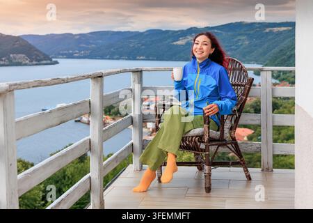 Lächelnde junge Frau entspannt sich in einem Korbstuhl auf einem Balkon mit einer Tasse und hat Blick auf ein Flusstal und eine Hügelstadt. Auszeit-Konzept. Stockfoto