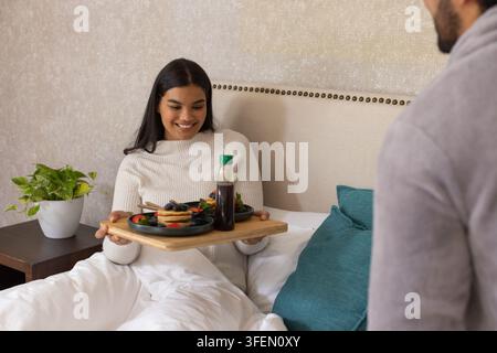 Afroamerikanisches Paar teilt das Frühstück im Bett mit Holztablett, Pfannkuchen, Sirup, Pullovern Stockfoto