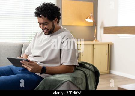 Mann sitzt auf hellgrauem Sofa im Wohnzimmer, hält ein Tablet und trägt eine schwarze Smartwatch Stockfoto