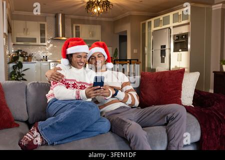 Paar sitzt auf grauem Sofa zu Hause, trägt Weihnachtsmützen und festliche Pullover, hält Smartphone Stockfoto