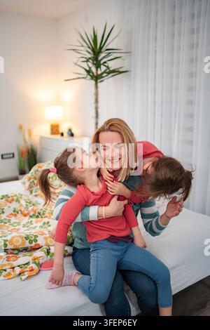 Glückliche Mutter umarmt und erhält Küsse von ihren verspielten jungen Töchtern im Bett Stockfoto