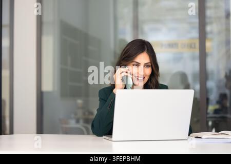 Geschäftsfrau telefoniert am Smartphone, während sie an einem Laptop arbeitet Stockfoto