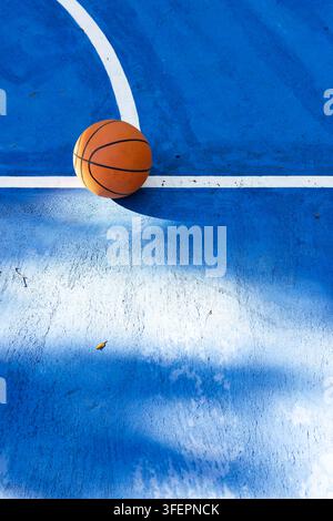Oranger Basketball, der auf blauem Betonplatz an der weißen Linienkreuzung ruht und einen langen Schatten wirft Stockfoto