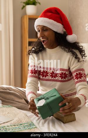 Afroamerikanische Frau sitzt auf dem Bett, trägt einen Weihnachtsmannhut, hält ein grünes Geschenk und eine kleine goldene Schachtel Stockfoto