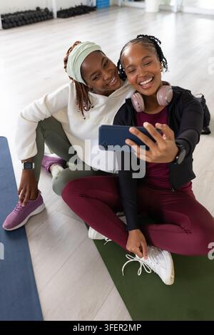 Zwei afroamerikanische Frauen sitzen auf Yogamatten und machen ein Smartphone-Selfie in einem hellen Fitnessstudio Stockfoto